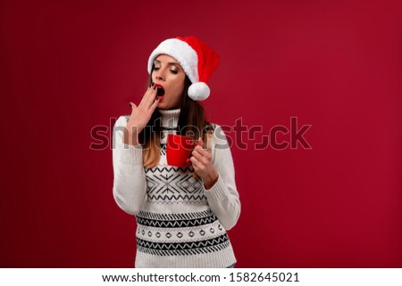 Close up portrait beautifiul caucasian woman in red Santa hat on red studio background. Christmas New Year holiday concept. Girl teeth smiling positive emotions with Red cup of coffee Lazy sleepyhead