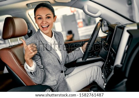 Attractive young businesswoman showing thumbs up is choosing new vehicle in car dealership.