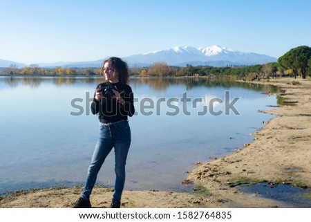 

Girl with curly hair takes pictures in a lake with the mountains in the background.Lake of Villeneuve-de-la-Raho (France) overlooking the Pyrenees and the Canigo. Concept: Landscape photography