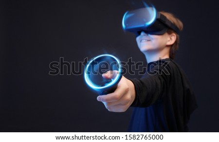 Young boy on dark background. Youngster using VR helmet with controllers in hands playing in video games and expresses happiness. Blue neon light.