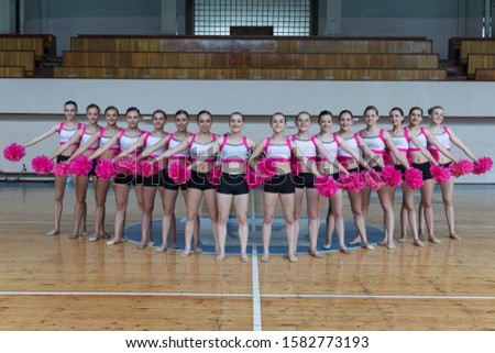 attractive young cheerleaders working out in sports club, group of cheerleaders with pom-poms in their hands, hands to the side, girls in black and pink suit with pompons on the background of the gym