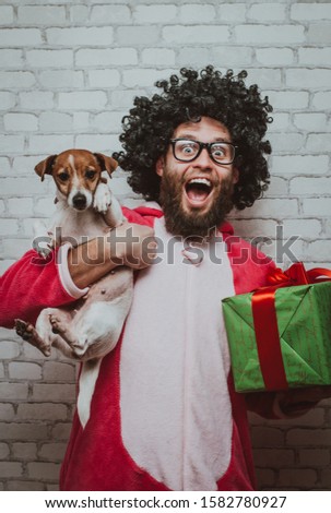 Merry Christmas or happy birthday! Cheerful bearded young man with curly hair in pink dragon pajamas with Christmas gift box and little jack rassell in hands. Crazy emotions. Pajamas party