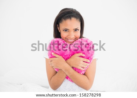 Delighted young dark haired model cuddling a heart-shaped pillow in bright bedroom