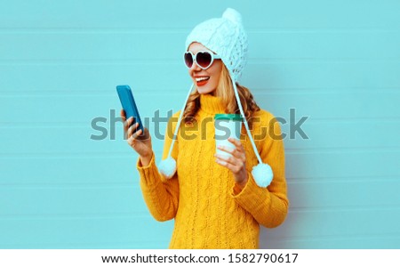 Winter portrait smiling young woman with phone and coffee cup wearing yellow knitted sweater, white hat with pom pom, heart shaped sunglasses on blue background