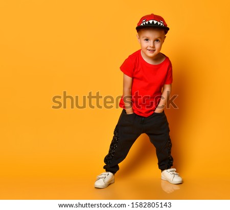 Little cute 4 year old boy in wide dark pants with pockets and a red T-shirt. A merry cap with shark teeth. Standing on a yellow background, put his hands in his pockets and looks at you