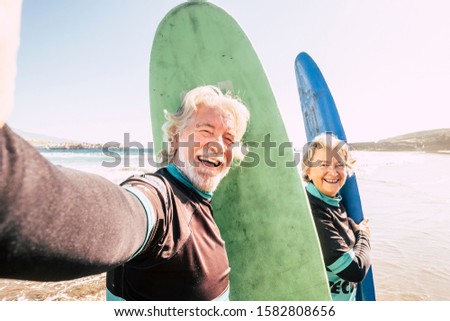 happy couple of seniors at the beach trying to go surf and having fun together - mature woman and man married taking a selfie with the wetsuits and surftables with sea or ocean at the background