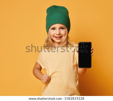 Smiling screaming little girl kid in green modern winter hat showing blank screen of new popular mobile phone on light yellow background