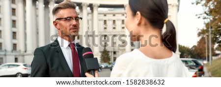 Cropped portrait of young woman conducting journalistic interview of cheerful politician. People making interview using equipment set at outdoor location. Horizontal shot. Selective focus on man
