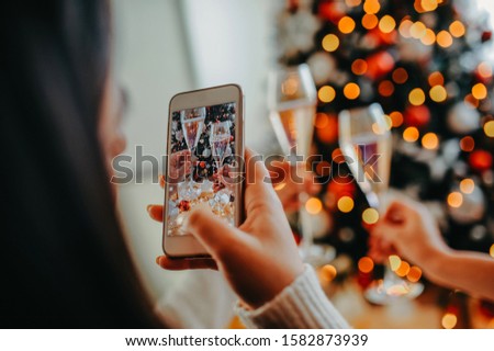 girl makes a photo on a white phone, two knocking glasses with champagne against the background of the Christmas tree in female hands