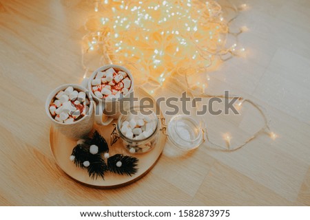 two white cups with a hot drink and white marshmallows, on a golden tray with a glass jar marshmallows, sprigs of a Christmas tree, on a wooden floor in a large number of lights from a garland