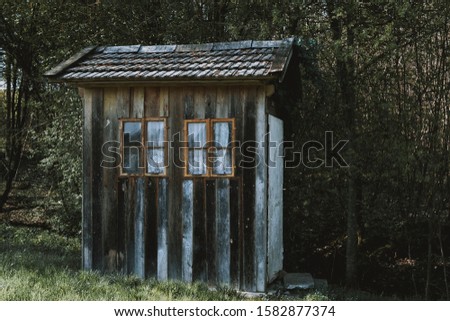 A small wooden cabin with brown windows with white curtains in a forest surrounded by trees