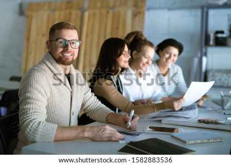Young successful designer drawing new fashion sketch on background of colleagues looking through papers