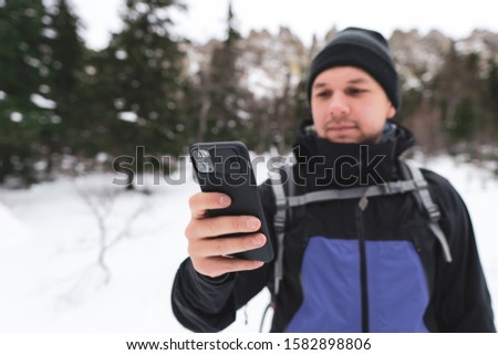 Young man hiker takes photo selfie on new smartphone; high steep snowy stone slopes of mountains and pine fir forest in snowy winter day on background; tourist look at the phone; mobile navigation
