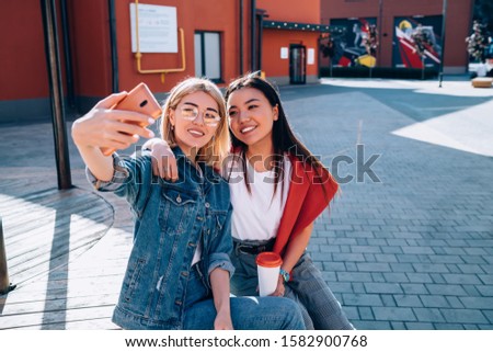 Young charming diverse females in casual apparel with cup of coffee making selfie with mobile phone in city in summer sunny day

