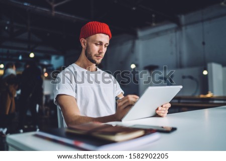 Concentrated young handsome guy sitting at table and drawing on big tablet with stylus pen working in spacious industrial office 