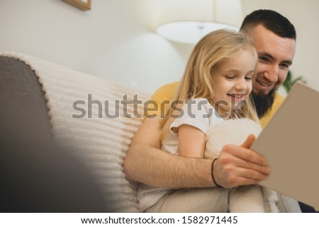Side view portrait of a lovely little girl reading a book with her father at home on the couch.