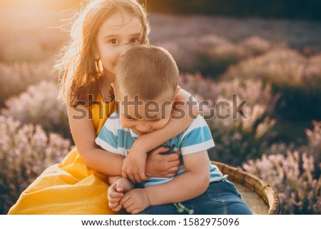 Cute little girl looking at camera while kissing her little brother and embracing him against sunset in a field of flowers.