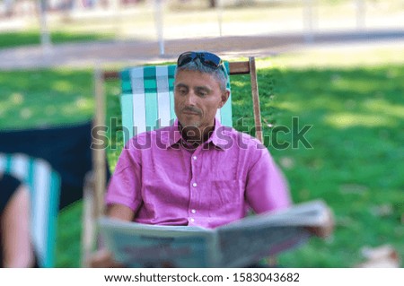 Relaxed man in a city park reading newspaper on a deck chair.