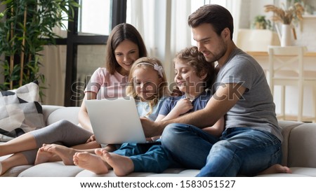 Married couple and little preschool kids using laptop ordering on-line, make purchase through e-commerce websites, choosing cartoon spend weekend together sitting on couch in cozy modern living room