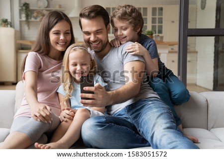 Cheerful family with little kids seated on sofa play new game on smartphone, take selfie photo having fun on-line using app, make video call talk with relatives, buying distantly e-commerce concept