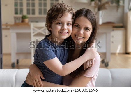 Son and mom sitting on couch at home and embracing celebrating mothers day looking at camera. New mum for adopted small kid boy, relative people elder sister and younger brother warm relations concept