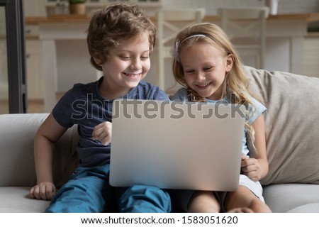 Two little kids siblings seated on sofa in living room holding on lap laptop play online game, watch cartoons spend weekend using electronic device, bad habit, generation addicted with gadgets concept