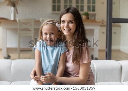 Attractive young babysitter and little kid girl hugging seated on couch look at camera, older younger sisters having warm relations and understanding, mother and daughter love and affectionate concept