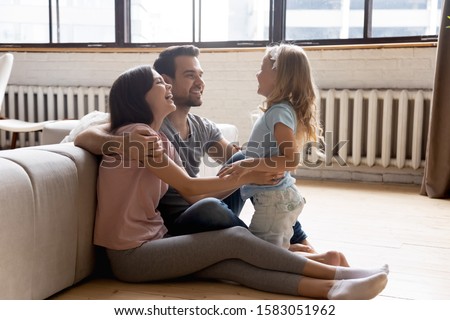 Adorable daughter spend time with loving mom and dad family in home clothes lean on couch sit on warm wooden floor with underfloor heating feel comfort having fun talking enjoy communication together Royalty-Free Stock Photo #1583051962