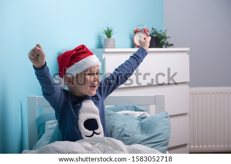 Funny child boy in Santa Clause hat and pajamas waking up in Christmas morning. Happy child sleep in white bed in child bedroom at home. Happy Xmas and New Year holiday! Christmas gift. 