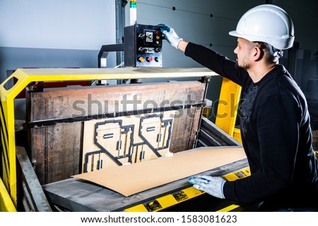 Turning on a punching machine. Worker in a hard hat in cardboard boxes factory working on paper die cutting machine. Royalty-Free Stock Photo #1583081623
