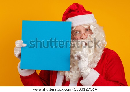Male actor in a costume of Santa Claus holds in his hands a blue sheet of paper for records and poses on a yellow background