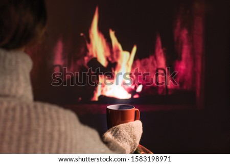 Woman in a winter sweater relaxes sitting near Christmas fireplace with a cup of hot drink and warming woolen blanket. Cozy atmosphere. Winter and Christmas holidays concept