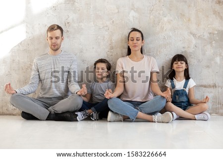 Mindful calm happy fit family of young parents and little kids sitting together on heated warm wooden floor in lotus positions, meditation, practicing breathing yoga exercises. Wellness hobby concept Royalty-Free Stock Photo #1583226664
