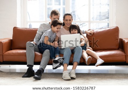 Happy family with two children having good time using laptop. Young smiling husband and wife with cute daughter and son sitting on sofa at home looking at computer screen and watching video.