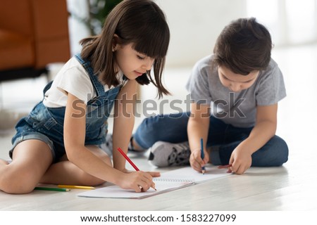 Brother and sister sitting on floor pencil drawing on paper. Happy smiling siblings children have fun and enjoy using fantasy on sofa background.