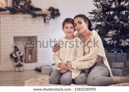 mother and son playing near the Christmas tree. Family celebratory Christmas evening. Mom and son on floor in white knitted sweaters hugging posing for photo