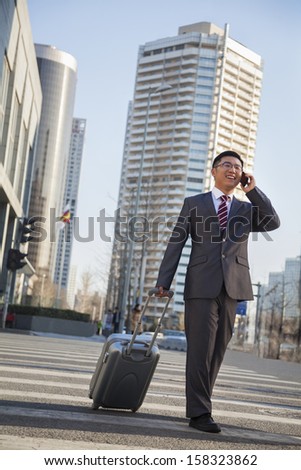 Smiling young Businessman walking down the street with luggage and on the phone