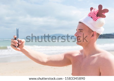Young man, handsome guy in Santa Claus hat on sea, beach making a selfie, taking a photo, picture of himself on his cell mobile smartphone, have a video call. Christmas winter vacation, happy New Year