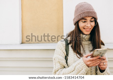 Attractive young woman standing outdoors against building's wall, playing on smart phone. Brunette female dressed in knitted sweater, hat, eyeglasses send messages from her device on the city street