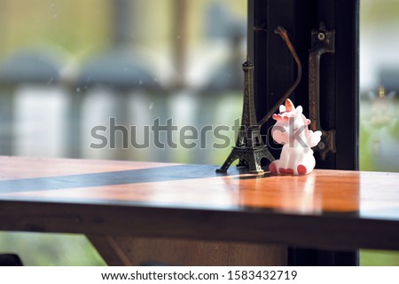 A unicorn doll put on wood table belong with an Eiffel Tower model put in front of a handle of window with sunshine on morning time.