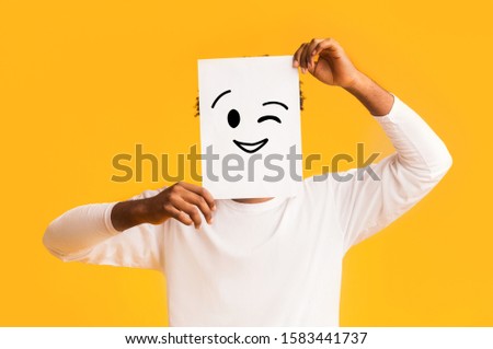 Black guy holding paper with positive emoji over orange background, hiding his real emotions