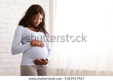 Peaceful pregnant black woman hugging her tummy next to window at home, free space Royalty-Free Stock Photo #1583457967