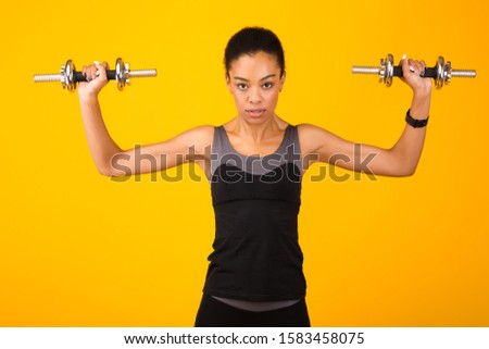 Determined Black Girl Exercising With Dumbbells Looking At Camera Standing Over Yellow Studio Background. Strength Workout Concept.