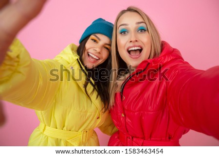 two attractive girl friends women taking selfie photo on pink background in colorful winter jacket of bright red and yellow color having fun together, warm coat sportswear fashion trend, crazy funny 