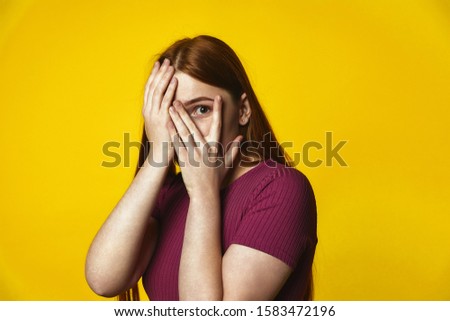 Front View of young redhead caucasian girl is covering face with hands and looking through the fingers on the yellow background dressed in pink t-shirt