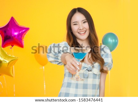 Portrait of Asian woman with long hair wearing Scottish dress, smiling for the camera at birthday party.Holds hand a cocktail blue drink.Standing on yellow background with colourful carnival balloons