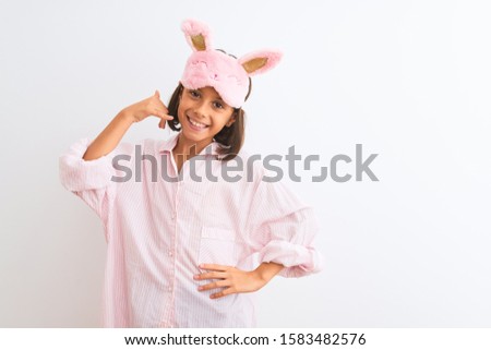 Beautiful child girl wearing sleep mask and pajama standing over isolated white background smiling doing phone gesture with hand and fingers like talking on the telephone. Communicating concepts.