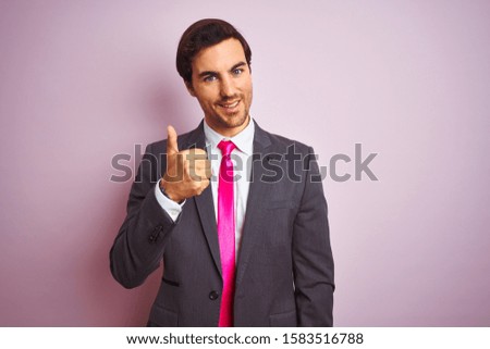 Young handsome businessman wearing suit and tie standing over isolated pink background doing happy thumbs up gesture with hand. Approving expression looking at the camera showing success.