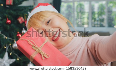 Asian women celebrate Christmas festival. Female teen wear Christmas hat relax happy holding Gift and using smartphone selfie with Christmas tree enjoy xmas winter holidays in living room at home.