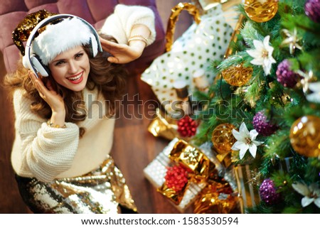 Upper view of smiling stylish housewife with long brunette hair in gold sequin skirt and white sweater listening to the music with headphones under decorated Christmas tree near present boxes.